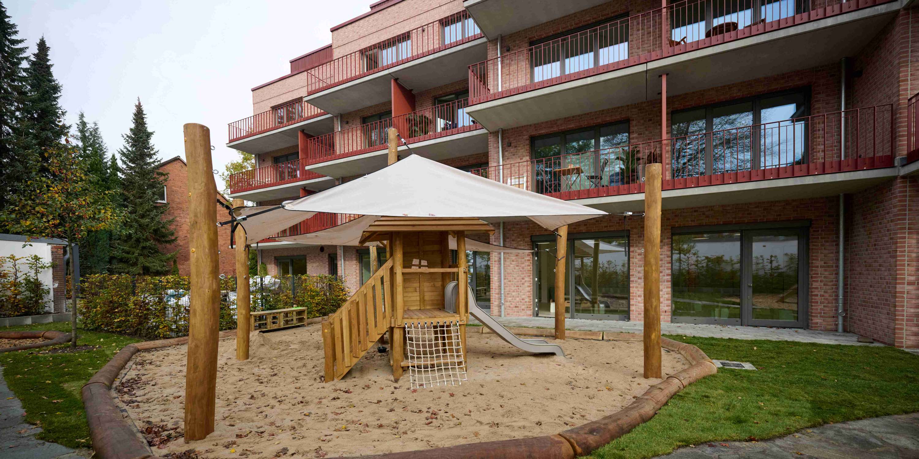 Ein kleiner Spielplatz im Innenhof eines modernen Wohngebäudes. Zu sehen ist ein hölzernes Spielhaus mit Rutsche und Kletternetz in einem Sandkasten, der von einem Sonnensegel überspannt wird. Im Hintergrund sind die großzügigen Balkone mit roten Geländern der angrenzenden Wohnanlage sichtbar.
