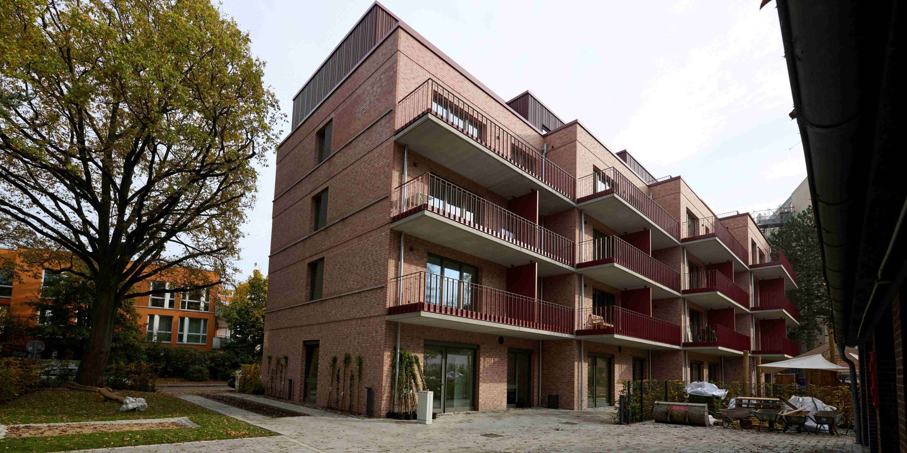 Die rückwärtige Fassade des Mehrfamilienhauses mit mehreren Reihen langer Balkone, die durch markante rote Metallgeländer auffallen. Der gepflasterte Innenhof ist teilweise von Bäumen gesäumt, und im Erdgeschoss sind bodentiefe Fenstertüren zu erkennen.