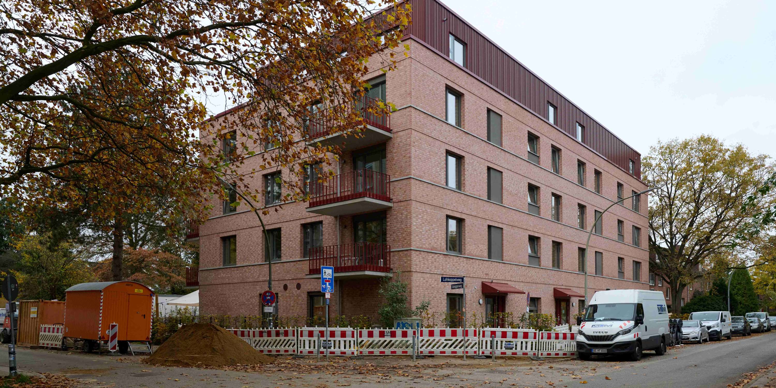 Ein moderner, vierstöckiger Klinkerbau an einer Straßenecke mit rötlich-beigen Ziegeln und einem abgesetzten Dachgeschoss in dunkler Metalloptik. Vor dem Gebäude befinden sich eine Baustelle mit Absperrgittern und ein großer Sandhaufen; herbstliche Bäume rahmen die Szene ein.