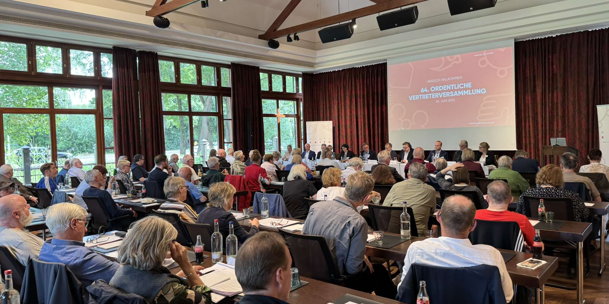 KAIFU-Vertreterversammlung: Ein Konferenzraum mit vielen Menschen an Tischen, die einer Leinwand zugewandt sind, auf der "64. ORDENTLICHE VERTRETERVERSAMMLUNG" steht.