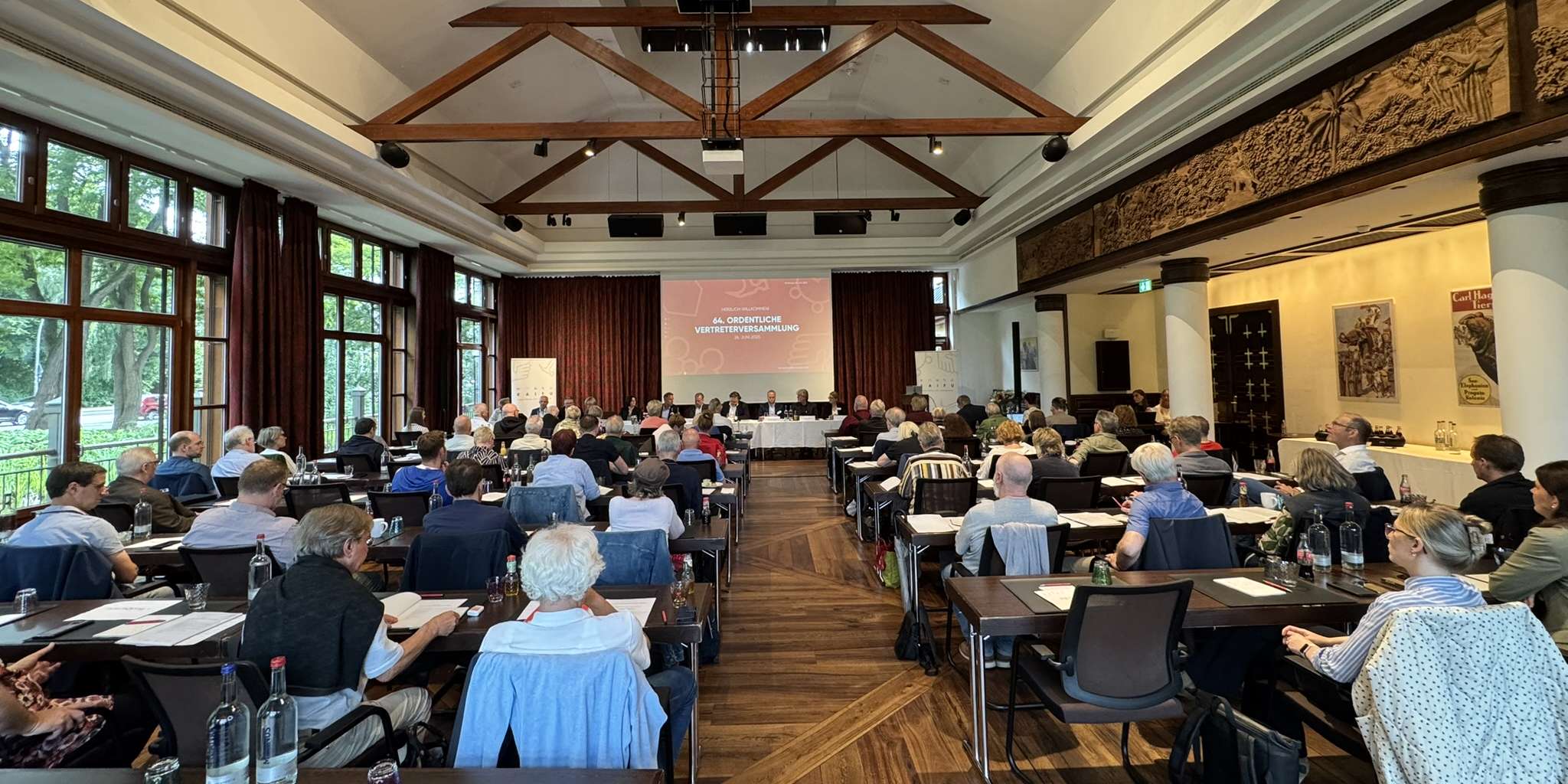 64, ordentliche Vertreterversammlung der KAIFU: Ein Konferenzraum mit vielen Menschen an Tischen, die einer Leinwand zugewandt sind.