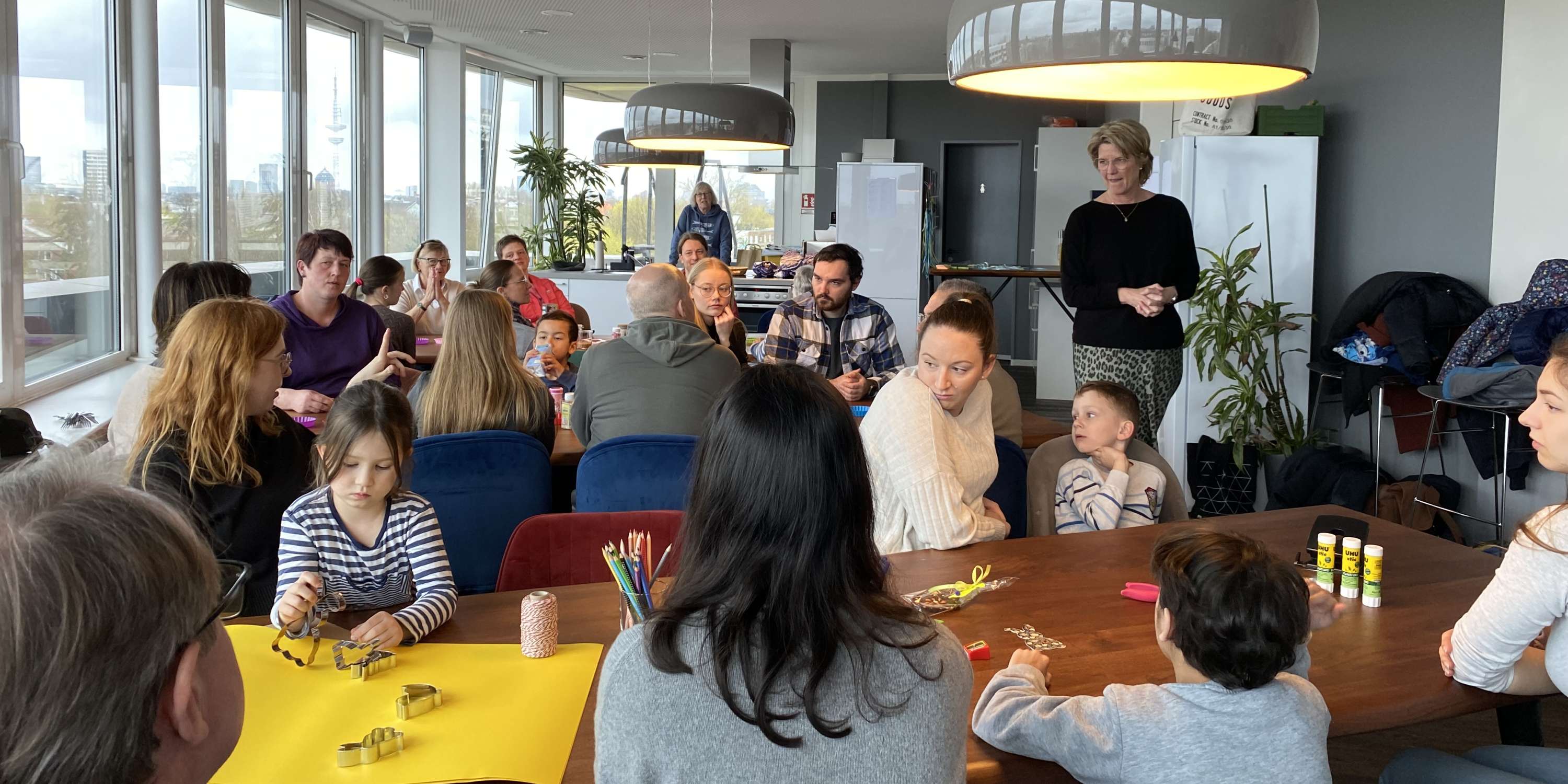 Eine Weitwinkelaufnahme eines belebten Gemeinschaftsraums, in dem viele Menschen unterschiedlichen Alters an langen Tischen gemeinsam basteln. Große, moderne Pendelleuchten erhellen den Raum. Auf den Tischen verteilen sich bunte Papierbögen, Stiftebecher und Bastelwerkzeuge. Durch die Fensterfront im Hintergrund blickt man auf eine Stadtlandschaft unter bewölktem Himmel.