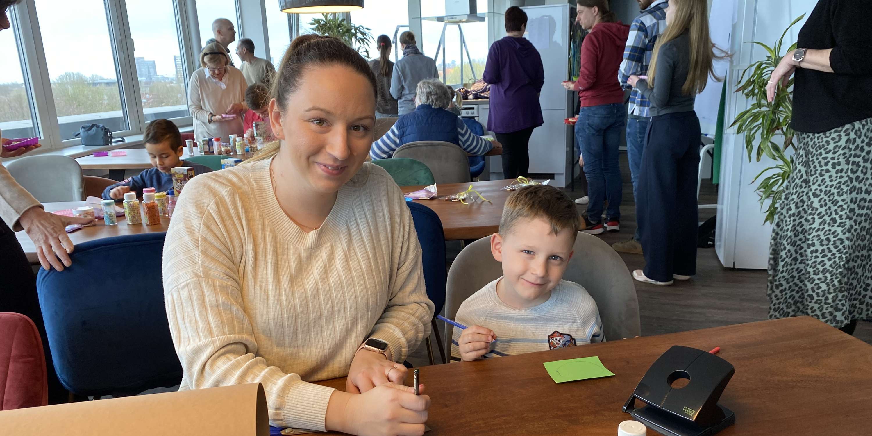 Eine Frau und ein kleiner Junge sitzen lächelnd an einem großen Holztisch und schauen in die Kamera. Vor ihnen liegen zahlreiche Bastelutensilien wie Klebestifte, Scheren, Motivlocher, bunte Stifte und eine große Rolle braunes Packpapier. Im Hintergrund sind weitere Personen in einem hellen, modern eingerichteten Raum mit großen Fensterfronten beim gemeinsamen Basteln zu sehen.