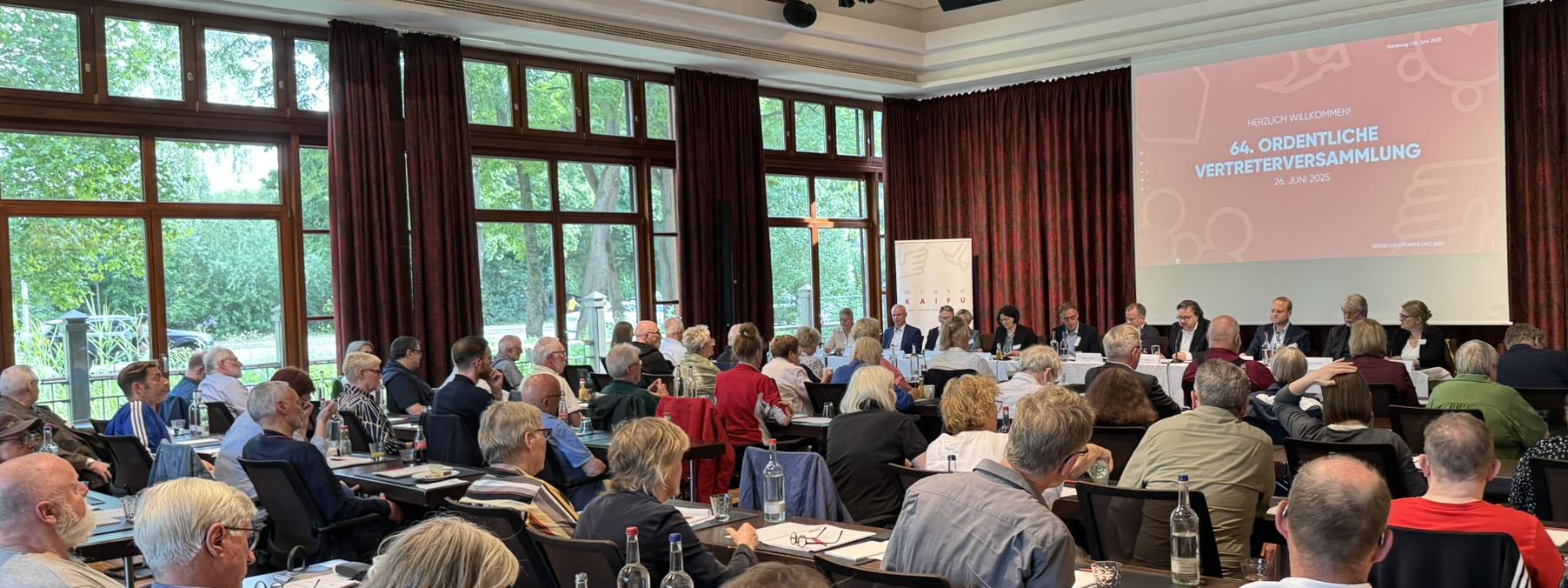 Ein Konferenzraum mit vielen Menschen an Tischen, die einer Leinwand zugewandt sind, auf der "64. ORDENTLICHE VERTRETERVERSAMMLUNG" steht.