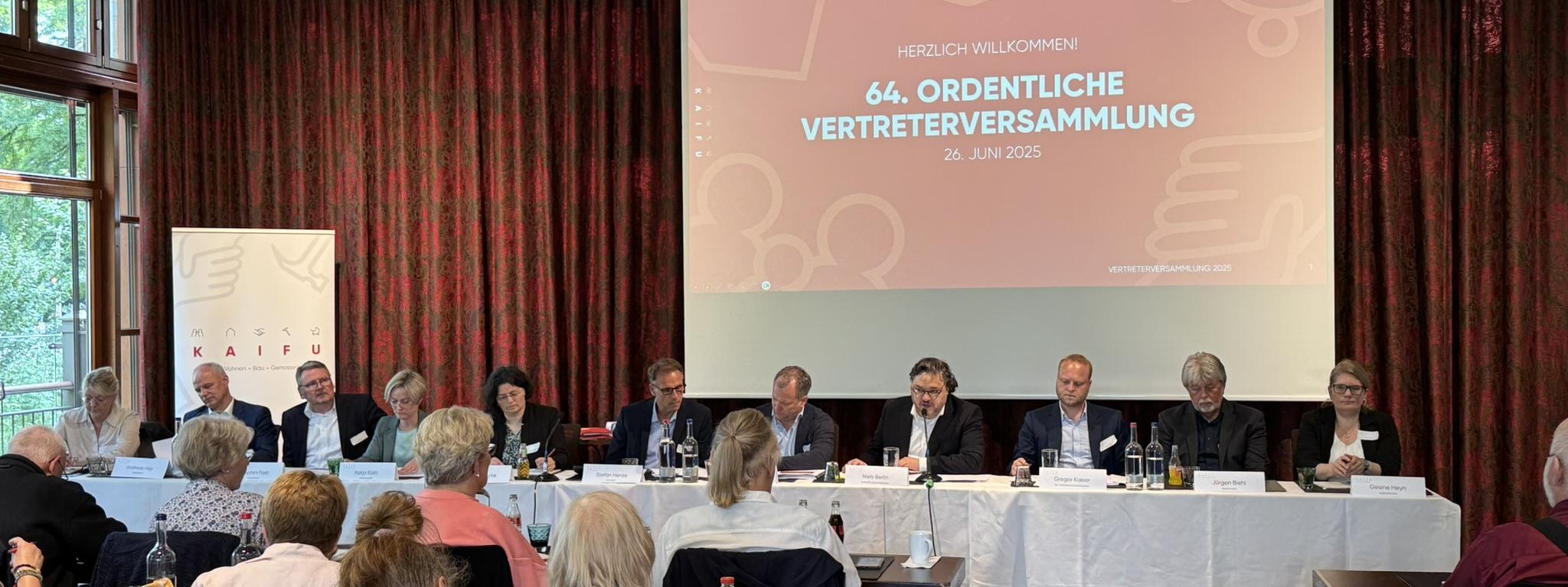  Ein Gruppenfoto zeigt eine Versammlung in einem großen Raum. Im Vordergrund sind die Köpfe von sitzenden Personen zu sehen, die in Reihen angeordnet sind. Dahinter befindet sich ein langer Tisch, an dem weitere Personen sitzen, die nach vorne blicken. Hinter diesem Tisch ist eine große Projektionsfläche aufgebaut, auf der in großen, roten Buchstaben "HERZLICH WILLKOMMEN! 63. ORDENTLICHE VERTRETERVERSAMMLUNG 20 JUNI 2024" steht. Der Raum ist mit roten Vorhängen und hölzernen Deckenbalken ausgestattet.