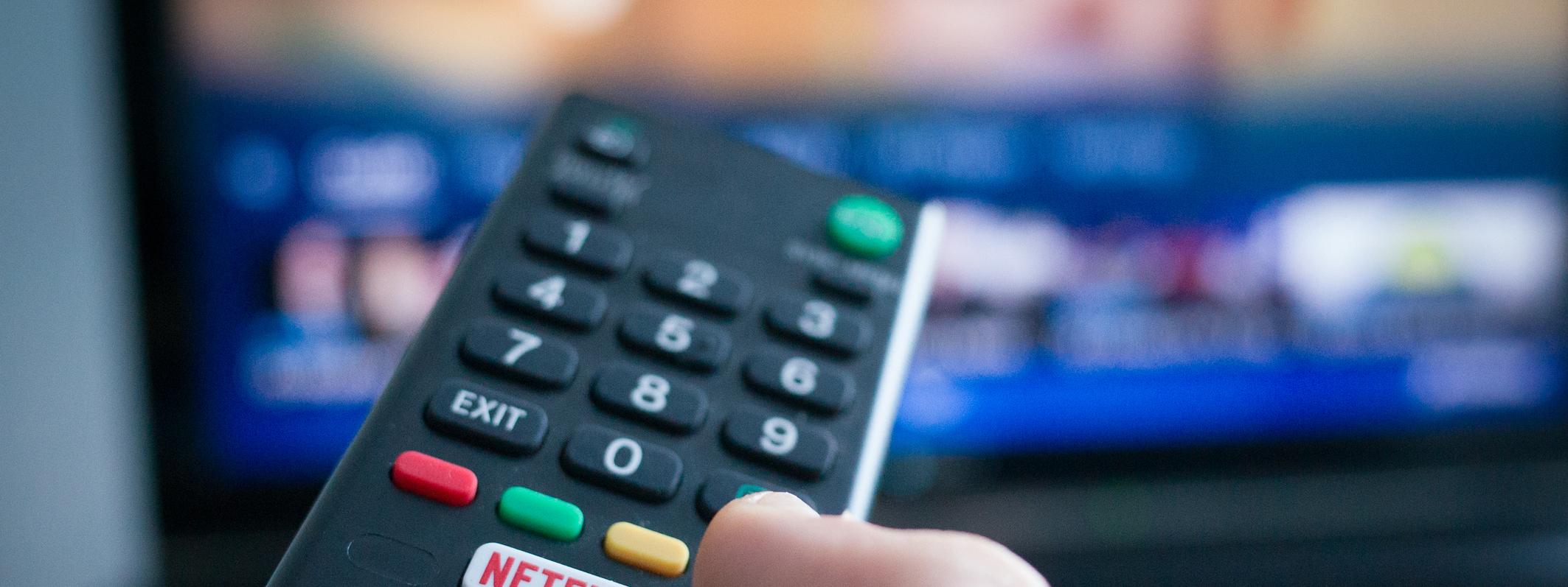 Eine Hand hält eine TV-Fernbedienung mit einer deutlich sichtbaren Netflix-Taste vor einem unscharfen Fernsehbildschirm.
